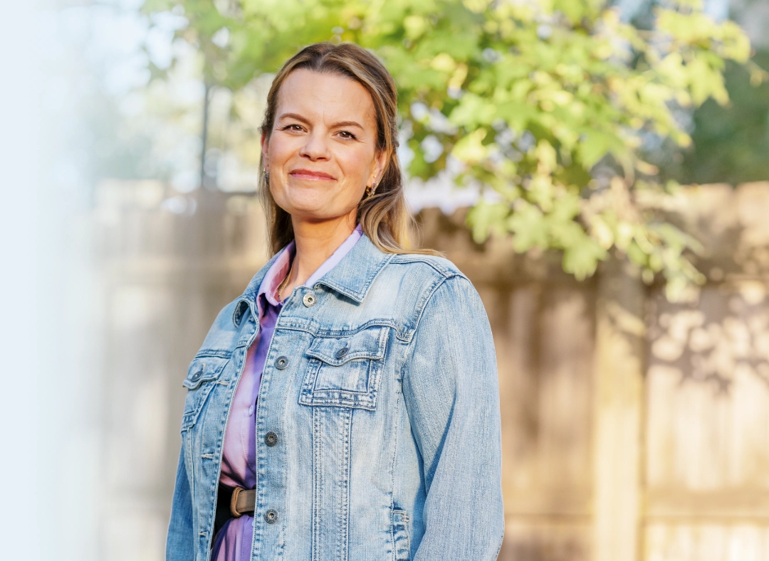 Image of a woman wearing a denim jacket over a purple shirt standing outdoors in front of the wooden fence