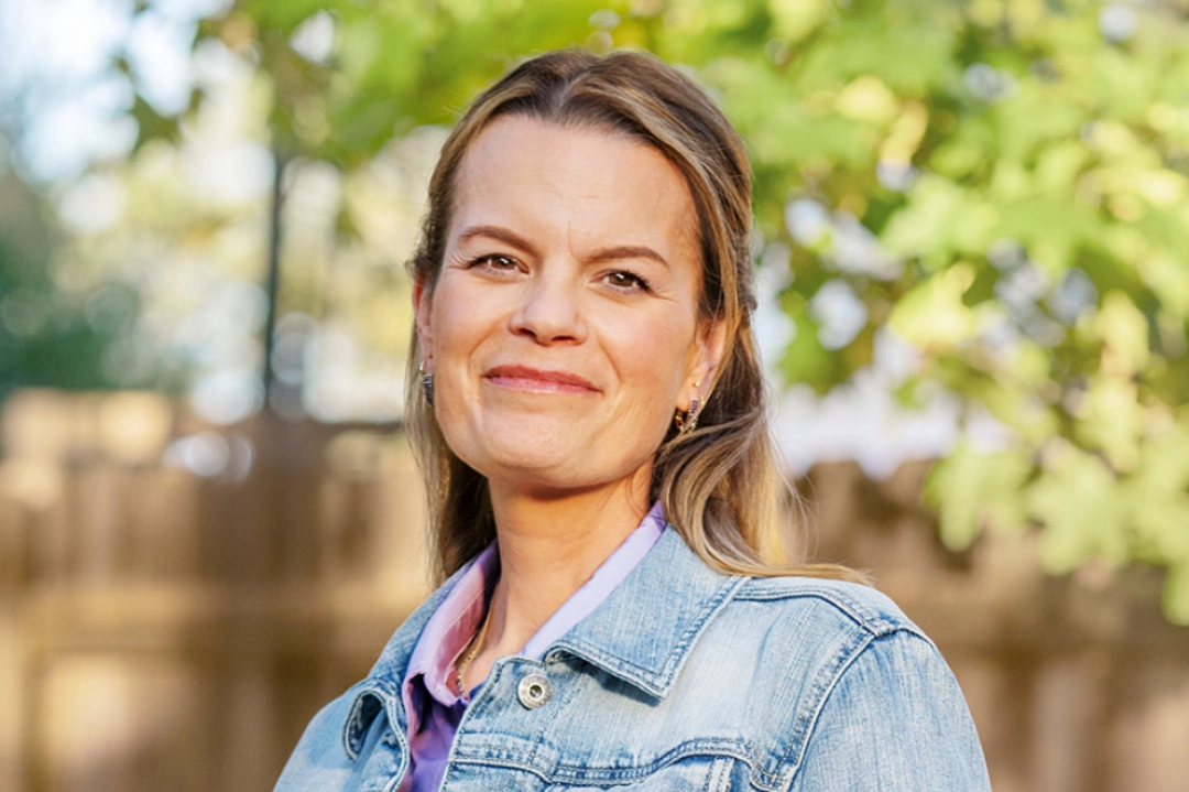 Image of a woman wearing a denim jacket over a purple shirt standing outdoors in front of the wooden fence