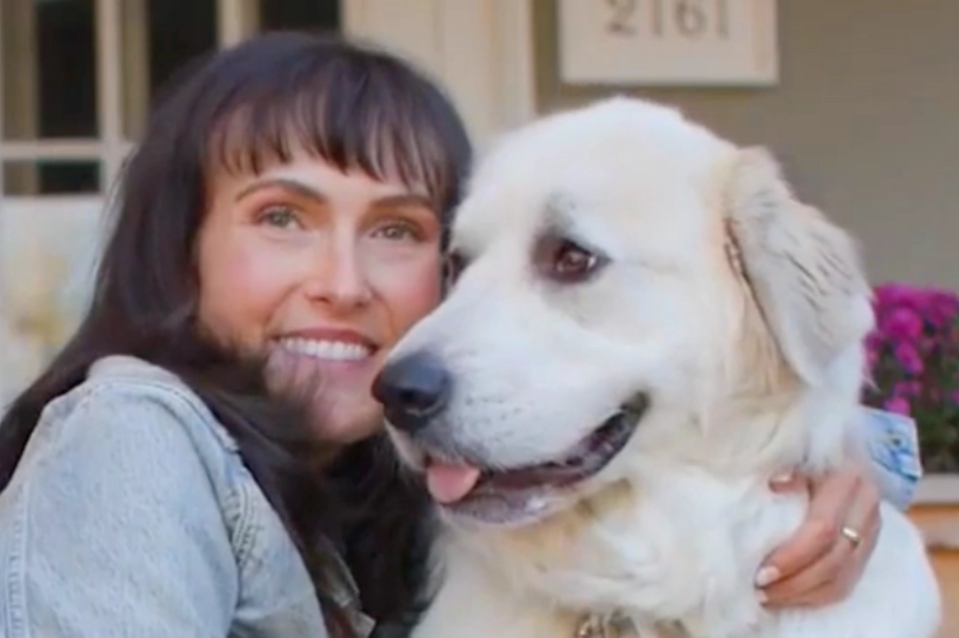 Image of woman in a denim jacket leaning her head against a white fluffy dog with its tongue slightly out, captured in a soft-focus outdoor setting